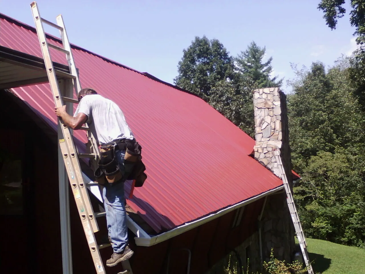 Roofer on a ladder beside a red metal roof and stone chimney in Mize
